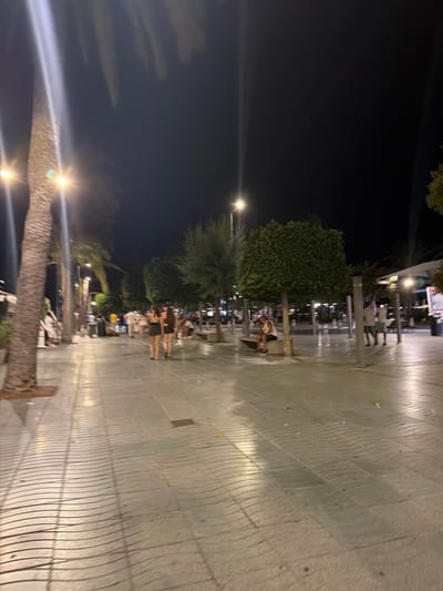 This image depicts a nighttime scene on a pedestrian walkway in Sant Antoni de Portmany, Spain. The walkway is paved with light-colored square tiles that have a distinctive wave-like pattern created by the shadows of surrounding elements and perhaps subtle variations in the paving. Several people are visible, walking or sitting. In the foreground, two women are walking away from the camera, and further down the walkway, a man is seated on a bench. Trees, including palm trees and other lush foliage, are interspersed along the walkway, illuminated by streetlights. The overall atmosphere is that of a lively evening promenade in a coastal town.