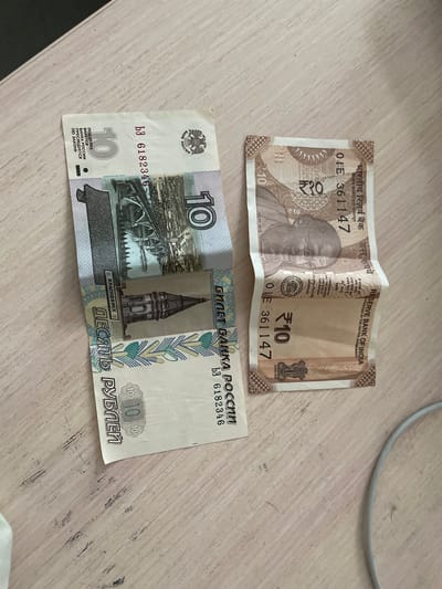 The image displays two folded banknotes on a light-colored, wood-grained surface. The banknote on the left is a Russian 10 ruble note, identifiable by the Cyrillic text "ДЕСЯТЬ РУБЛЕЙ" and the image of the Krasnoyarsk Bridge. The banknote on the right is an Indian 10 rupee note, featuring Mahatma Gandhi's portrait and the text "RESERVE BANK OF INDIA." The lighting suggests an indoor environment, likely a desk or table. There are no people or actions depicted. The image does not provide any information about the location in the city of Черниговка, Russia.