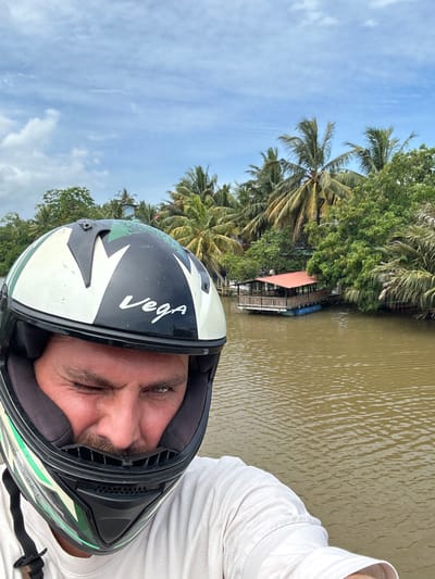 A man wearing a black, white, and green Vega motorcycle helmet is taking a selfie in front of a body of water. He is winking with his left eye. Behind him, lush green palm trees line the murky brown water, and a floating structure with a red roof is visible on the water's edge. The sky is overcast with scattered white clouds. The scene appears to be in Kadewatta, Sri Lanka, known for its tropical landscapes and waterways.