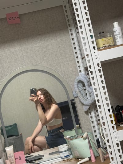 The image captures a woman taking a mirror selfie in what appears to be a dressing area or a bedroom in Dongducheon-si, South Korea. The woman, with wavy brown hair and wearing a white bralette and denim shorts, is seated, holding a smartphone to take the photo. In the foreground, a large circular mirror reflects her and the surrounding environment.
To the right of the mirror, a tall white metal shelving unit is visible, stocked with various personal care items. These include bottles of what appear to be lotions or toners, a jar of cotton swabs, and other cosmetic products. A light blue, plush headband with ear-like accents is hanging from the shelving unit.
On a surface in front of the mirror, several other items are placed, including makeup bags, tubes of cosmetics, and small containers. A pink sticky note with Korean writing is affixed to the textured wall above and to the left of the mirror. The overall impression is of a personal space, likely a bedroom, where the woman is getting ready.
