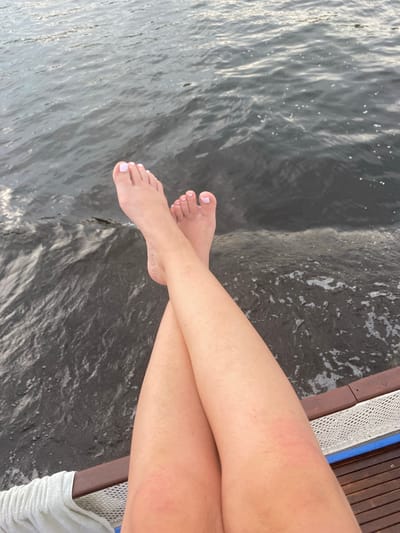A close-up, aerial view shows a person's legs and feet crossed over each other, resting on the edge of what appears to be a dock or boat. The water is dark and slightly choppy, with some ripples and white foam visible. The person's toenails are painted a light pink color. The legs are tanned and smooth, with a slight redness on the right thigh. The background is composed entirely of water. The location is not specified.