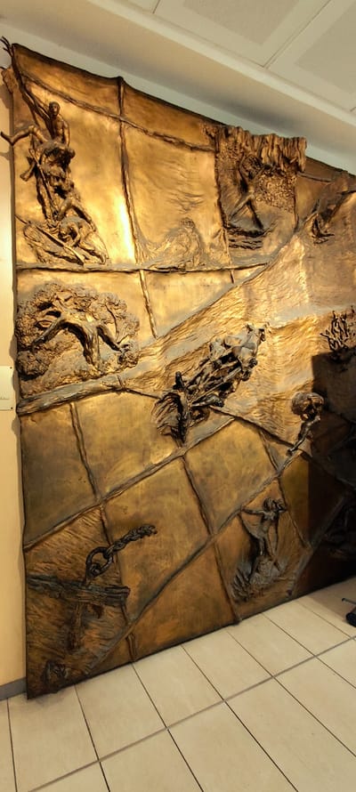This is a detailed image of a large, bronze relief sculpture mounted on a wall. The sculpture depicts a chaotic and dynamic scene with multiple figures and elements, rendered in a textured, three-dimensional style. The bronze has a golden patina, highlighting the raised details. The sculpture appears to be divided into sections by grid-like lines, adding to its structured yet complex composition.
In the upper left section, figures are shown in strained or acrobatic poses, one appearing to climb or pull on a draped element. Below this, a detailed representation of a tree with gnarled branches is prominent. In the upper right, a figure is visible on a raised area, with a small architectural structure resembling a temple on a distant hill. The central part of the sculpture shows a figure seemingly in motion, possibly riding or being carried. Further down, a segment displays a dark, twisted metal chain linked to a fixture. In the lower right, two figures are depicted, one in a more dynamic, dancing pose, and another standing on a rounded form. The overall impression is one of movement, struggle, and fragmented narratives.
The sculpture is installed on a light-colored wall, with a tiled floor visible in the foreground. The lighting creates strong highlights and shadows, emphasizing the sculptural form. There is a small informational plaque on the wall to the left of the sculpture, partially obscured.
This sculpture is identified as "Il Mito di Minosse" byettore F. by Ettore Ferrari, located in the Palazzo Comunale (Town Hall) in Marsala, Italy. The work is an allegorical representation of the myth of Minos, with various figures and symbols alluding to the legendary king of Crete and his labyrinth.