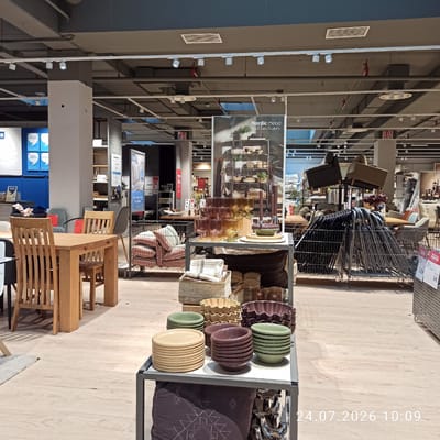 The image shows the interior of a furniture and home goods store. In the foreground, there is a wooden dining table with chairs. To the right, a display shelf holds various colored bowls and plates. Behind this, a metal cage contains stacked cushions. Further back, more furniture, including sofas and chairs, are displayed. Several posters and promotional materials are visible on walls and stands. The environment appears to be a retail space with a polished wooden floor and exposed ceiling infrastructure with track lighting. The time stamp "24.07.2026 10:09" is overlaid on the image. This scene is located in the IKEA store in Modugno, Italy.