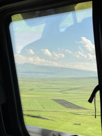 This media file is a photograph taken from inside a vehicle, likely a car or van, looking out through the windshield. The primary subject is a vast, green landscape divided into rectangular fields, suggesting agricultural land. The fields are predominantly a vibrant green, with some areas appearing darker, possibly indicating different crops or soil conditions. One large, dark rectangular patch in the middle of the green fields stands out.
The sky above is blue with scattered white, fluffy clouds. The perspective indicates that the photograph was taken from a moving vehicle, as suggested by the reflections on the windshield, which show parts of the vehicle's interior, including a dark frame around the windshield and a black strap or lever. These reflections create a layered effect, obscuring some of the view of the landscape.
The scene appears to be set during the daytime, with bright sunlight illuminating the landscape. The time of day seems to be late morning or early afternoon, given the brightness and the angle of the sun. The weather appears clear and sunny, with fair-weather clouds. The landscape suggests a rural or semi-rural location, characterized by open fields and a distant horizon.
There are no people or explicit events depicted. The overall impression is one of a scenic drive through the countryside. The image is oriented vertically, suggesting it might have been taken while tilting the camera or during a turn. There is no visible text within the image. The location context provided is Tsilkar, Armenia, which aligns with the appearance of the agricultural landscape.