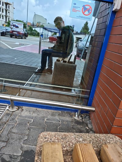 A young man wearing sunglasses and a green hooded jacket sits on a concrete bench outside a store in Mława, Poland. He is looking at his phone. The store has a glass facade with a blue frame. A red brick wall is visible to the right of the entrance. In the reflection on the glass, a sign reads "W NASZYM SKLEPIE OBOWIĄZUJE ZAKAZ WPROWADZANIA ZWIERZĄT WYJĄTKIEM SĄ PSY ASYSTUJĄCE OSOBOM Z NIEPEŁNOSPRAWNOŚCIAMI", which translates to "In our store, the bringing of animals is prohibited, except for assistance dogs for people with disabilities." Several cars and shopping carts are visible in the background, as well as a construction site. The ground is paved with interlocking concrete blocks.
