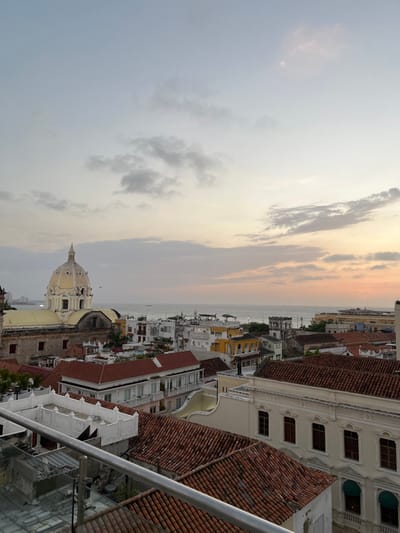 An aerial view shows a cityscape at dusk. The sky is a mix of blues and yellows, indicating sunset. Below, buildings with red-tiled roofs and white walls line the streets. A domed church stands out among the structures. The scene looks out over a body of water.
