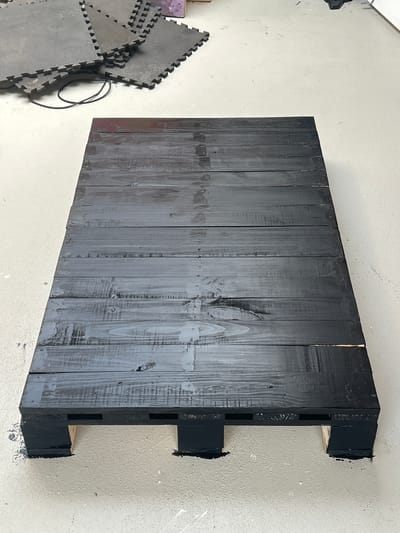 In Lisse, Netherlands, a black wooden pallet lies on a concrete floor. The pallet is made of planks and has visible wood grain. To the left of the pallet, there is a pile of interlocking foam mats. The floor is stained in some areas.