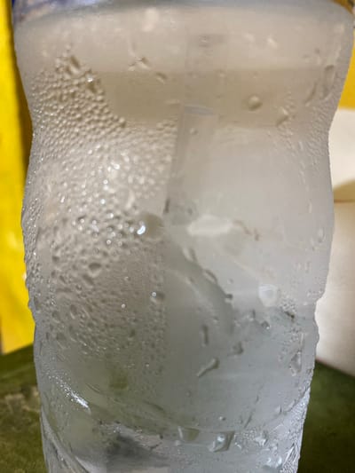The image shows a plastic bottle covered in condensation droplets, likely from a cold beverage. The bottle appears to be slightly deformed and contains a liquid inside. The background is blurred and includes yellow and green elements. The location is unknown, with no information in the image to indicate it is in Tanjongkapai, Indonesia. There are no people visible in the image.