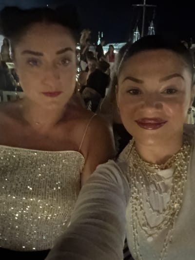 The media file is a close-up, sideways selfie of two young women. The woman in the foreground, on the right, is wearing a sparkly, off-the-shoulder top. She has dark hair and is wearing dark lipstick. Her head is tilted back, and she is looking up and to her left. The woman in the background, on the left, has lighter hair and is wearing a white top with beading around the neckline. She is also looking away from the camera. The background is dark, suggesting it is nighttime. There appear to be some lights and structures in the background, possibly indicating an outdoor event or gathering. The overall mood is celebratory and intimate, as is common in selfies. No text is visible in the image. The location context of Naousa, Greece, might suggest a festival or a nightlife scene.