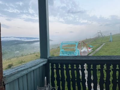 The image shows a view from a wooden balcony overlooking a misty mountainous landscape in Nikortsminda, Georgia. In the foreground, the dark wooden railing of the balcony is visible, with a patterned cutout design. Beyond the balcony, a blue swing set and a wooden deck are present on a grassy slope. Further in the distance, rolling hills covered in lush green vegetation are shrouded in a light mist, with glimpses of a village nestled among the trees. The sky is overcast with soft, diffused light. There are no people or animals visible in the image. The overall atmosphere is serene and tranquil.