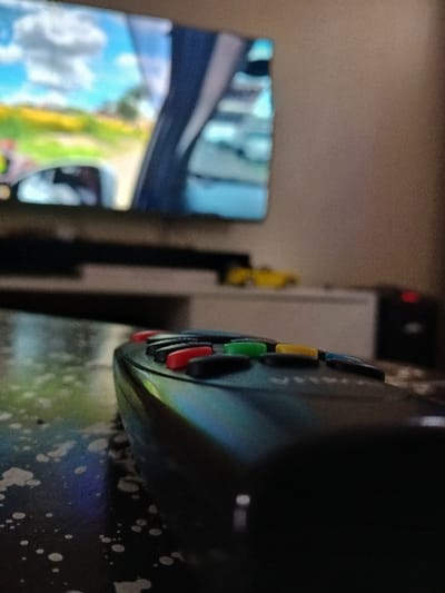 The image shows a black TV remote control lying on a dark surface. The remote control is in focus, while the background is blurred. The remote control has several buttons, including four colored buttons: red, green, yellow and blue. It is in a room in Gitaru, Kenya (latitude: -1.232, longitude: 36.676). The image was taken in a room, with a television in the background. The image is captured from a low angle, with the remote control in the foreground. The image was taken indoors, with a smooth, shiny surface.
