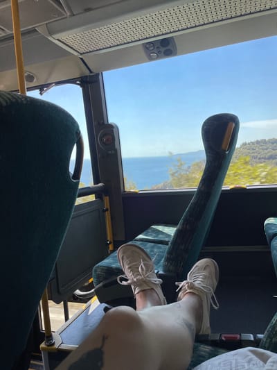 This media file captures a low-angle perspective from inside a bus, looking out towards a bright, sunny sky and a scenic landscape. The subjects are a person's legs and feet, clad in light-colored sneakers, resting on the bus floor. Their legs are positioned as if they are comfortably seated, with one leg crossed over the other.
The scene outside the bus window reveals a body of water, likely a bay or sea, with mountains or hills covered in lush greenery in the background. The sunlight suggests it is daytime, possibly late morning or early afternoon, and the clear sky indicates fair weather. The interior of the bus is visible, with green upholstered seats and a yellow handrail. There are also components of the bus's interior structure, including a ventilation grate and some electronic controls on the dashboard.
The overall impression is one of travel and relaxation. The person's relaxed posture and the view of the picturesque landscape evoke a sense of ease and enjoyment during the journey. The location context provided, Perazića Do, Montenegro, helps to identify the specific scenery outside the bus as a coastal area in that region. No visible text is present in the media file.