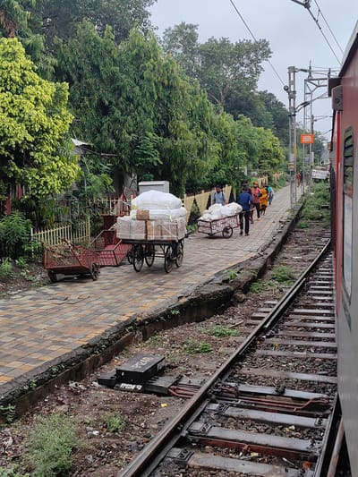 The image depicts a scene at the Bhopal Junction railway station in Bhopal, India. In the foreground, a train track runs parallel to a paved platform. The platform is adorned with patterned tiles and bordered by a low concrete wall. On the platform, several carts are visible. One cart, laden with white sacks and boxes wrapped in plastic, is being pushed by a man in a blue shirt. Another smaller, empty cart sits nearby. In the background, a line of trees and foliage forms a green backdrop to the station. Several people are walking along the platform, some carrying bags. A red train car is partially visible on the right side of the frame, indicating that the photo was taken from within a train or from a close vantage point. The sky appears overcast, suggesting a cloudy or possibly rainy day. Electrical wires and poles associated with the railway system are also present. The overall impression is a busy railway station environment with people and goods being transported.