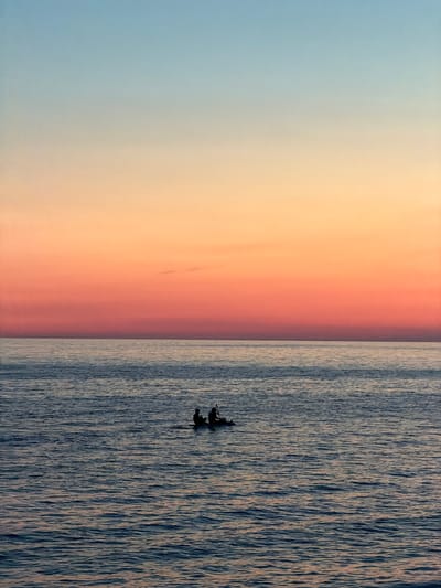 Two individuals are seated in a small boat on the calm sea, their silhouettes dark against the vibrant sunset sky. The sky transitions from a pale blue at the top to shades of orange and pink, indicating a sunset or sunrise. The water is a deep blue, with subtle ripples reflecting the colors of the sky. The scene is set on the coast of Perazića Do, Montenegro.
