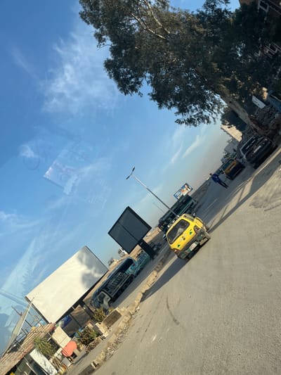 An angled perspective captures a street scene with a mix of vehicles and urban elements. A bright yellow car and other vehicles of darker hues line the roadside. A large tree stands to the side, contrasting with the clear blue sky reflecting off a nearby glass surface, mirroring clouds and possibly an advertisement. Street lamps, building structures, and various roadside objects contribute to the bustling yet somewhat tilted cityscape.
