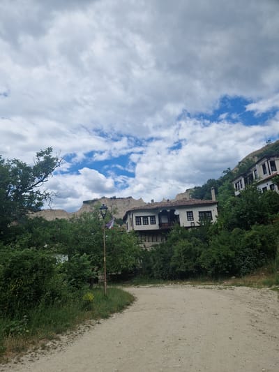 A dirt road curves through lush green foliage and leads towards traditional Bulgarian houses. The houses, with their white walls and dark-tiled roofs, are nestled into a hillside, suggesting they are part of the historic town of Melnik. The background features dramatic sandstone rock formations, characteristic of the Melnik region, under a sky filled with dynamic clouds. A street lamp stands prominently in the foreground, adorned with a Bulgarian flag, adding a touch of local identity to the scene. The overall impression is one of a charming, historical setting in a picturesque natural environment.