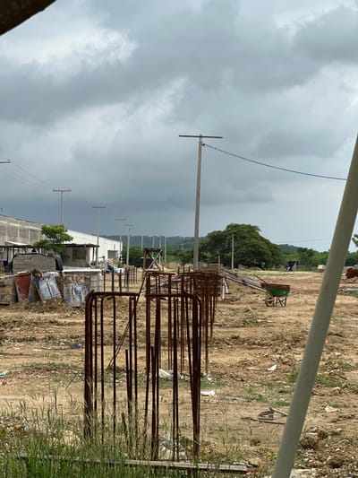 The image depicts a construction site under a cloudy sky in Juan Mina, Colombia. In the foreground, vertical rusty rebar posts are lined up, partially obscured by tall green grass. To the left, several large, weathered barrels, some with blue and white markings, are stacked on a dirt mound. Behind the barrels, a long, low-slung building with a corrugated metal roof and white siding is visible. Several utility poles with wires stretch across the scene. Further into the background, a green wheelbarrow with a wooden handle is parked on the uneven, dirt terrain. A large, leafy green tree stands prominently in the middle ground. Two individuals are standing in the distance near a small structure. The overall environment is characterized by construction materials, raw earth, and a slightly overcast atmosphere, typical of a developing area.