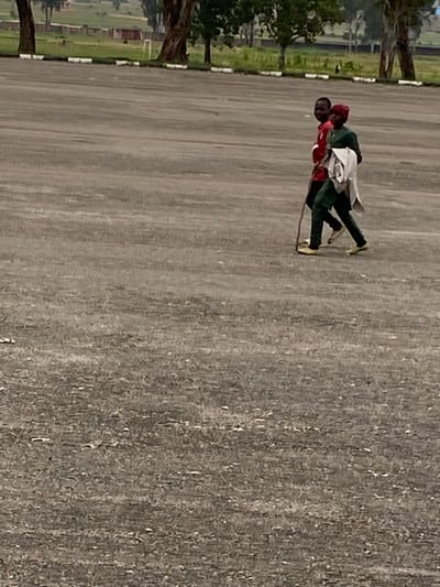 In Zawan, Nigeria, two individuals are walking across a paved outdoor area. The person in front is wearing a green outfit with a red head covering and carrying a folded white cloth over their left shoulder. They are holding a wooden stick in their right hand. The person behind them is wearing a red shirt and appears to be looking towards the viewer. They are walking side-by-side. The ground is a rough, grey asphalt. In the background, there is a strip of green grass with several trees, and beyond that, a fence and some buildings. The sky is overcast.