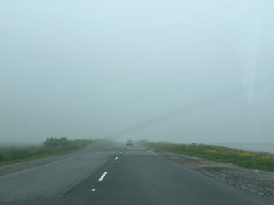 The image depicts a road in a foggy environment. The road is paved and has white lane markings. On either side of the road, there is vegetation, including grass and some bushes or trees. In the distance, a car is visible traveling on the road, partially obscured by the fog. The sky is overcast and appears very hazy, contributing to the overall low visibility. There are no people visible in the image. The scene suggests a rural or semi-rural setting with limited infrastructure. The specific location in the city of Sovetskoe, Russia, cannot be determined from the visual information alone.
