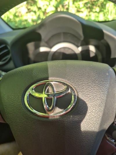 The image shows a close-up of a Toyota steering wheel, with the Toyota logo prominently displayed in the center. The steering wheel appears to be made of dark-colored, textured material, possibly leather. Sunlight is casting a shadow across the steering wheel, creating a gradient of light and dark. In the background, through the windshield, a blurry view of green foliage is visible, suggesting the vehicle is parked or driving outdoors. The dashboard and instrument cluster are partially visible behind the steering wheel, but they are out of focus. There are no people or specific landmarks visible that would indicate the location in Trier, Germany.