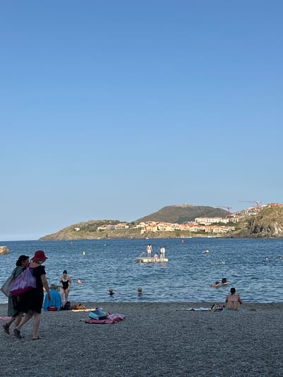 The image depicts a view of the beach and the sea in Collioure, France, with the town's hillside architecture and a mountain in the background. Several people are visible. In the foreground, three women are walking on a pebble beach. One woman wears a green dress and carries a striped tote bag, another wears a black dress and a red hat, and the third is out of frame. Behind them, several beach towels and bags are scattered on the sand. A woman in a black swimsuit is standing near the water, and another person is sitting on a towel.

In the water, numerous people are swimming or wading. A yellow buoy is visible, as is a floating platform with several individuals on it. Further out, more swimmers can be seen. The sea is calm and blue, and the sky is clear and cloudless. In the distance, the town of Collioure cascades down a rocky hillside, with buildings in shades of white and terracotta. Construction cranes are present on the upper slopes, indicating ongoing development. The overall scene suggests a warm, sunny day at a popular coastal resort.