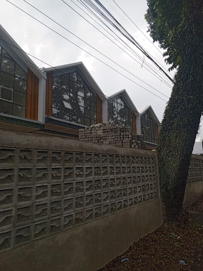 The image displays an outdoor scene in Qorday, Kazakhstan, under an overcast sky. In the foreground, a concrete retaining wall runs horizontally across the frame, topped with a section of interlocking grey concrete breeze blocks featuring a repetitive square pattern. The ground directly in front of the wall and to the right is covered with dark brown organic material, primarily fallen leaves, interspersed with scattered small pieces of white and blue plastic debris. On the far right, a large tree with a thick trunk is visible, heavily covered in dense green vines.
Behind the retaining wall and beneath the buildings, a substantial stack of light-grey rectangular building blocks is present. The midground and background are dominated by a series of modern architectural structures. These buildings feature prominent gabled or "sawtooth" roofs, characterized by light-colored eaves. The facades primarily consist of large, dark-tinted glass windows that reflect portions of the sky and the surrounding tree branches. Vertical sections of light-brown wooden paneling separate the window units. One window on the central building is noticeably ajar, tilted open from the bottom. Multiple black utility lines are visible stretching horizontally across the upper third of the frame against the bright, cloudy sky. No people, active movements, or interactions are depicted in the scene.