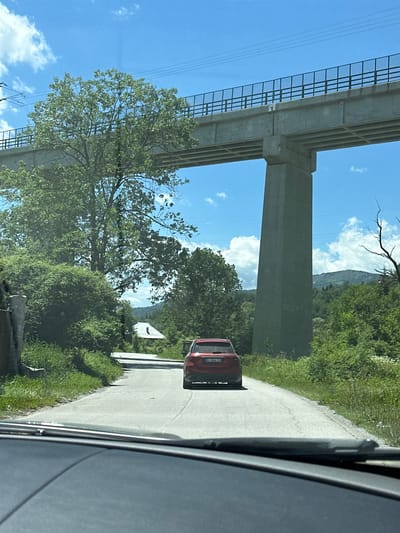 A red SUV is driving down a paved road in Mušovića Rijeka, Montenegro. The SUV is in the middle of the road, and the view is from the perspective of someone in another car behind it. To the left of the road, there is a large tree and dense green foliage. To the right, there is more green foliage and a large concrete bridge support pillar. Above the road and trees, a concrete bridge with a metal railing spans across the scene. The sky is blue with scattered white clouds. In the distance, there are rolling green hills.