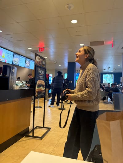 The image captures an indoor scene within a university cafeteria or food service area at Brandeis University in Waltham, Massachusetts, United States.
In the right foreground, a young adult female with blonde hair pulled into a ponytail, wearing a light tan cable-knit sweater and dark pants, stands smiling widely and looking upwards and to the right. She holds a black camera with a neck strap. To her immediate right, a brown paper bag rests on a white surface.
The mid-ground displays a food service counter with a dark top and light wooden base. Behind this counter, multiple digital menu boards are visible, advertising items such as "RISE & SHINE BOWLS" and "FULLY LOADED." A male individual in a dark long-sleeved shirt stands at the counter, appearing to be engaged in an transaction. A freestanding black sign with an advertisement for a "FRESH START" burger is prominent, accompanied by a smaller sign for a "TREAT" cinnamon roll. A queue line, delineated by black stanchions with retractable belts, extends into the background, where another male in a dark vest stands.
The background features additional patrons seated at tables in a dining area. Large windows on the right wall offer a view of outdoor trees and greenery. The ceiling is a white drop-tile design, equipped with recessed lighting and red "EXIT" signs. The floor is covered in light-colored square tiles. A blue and grey trash/recycling bin is partially visible on the far right.