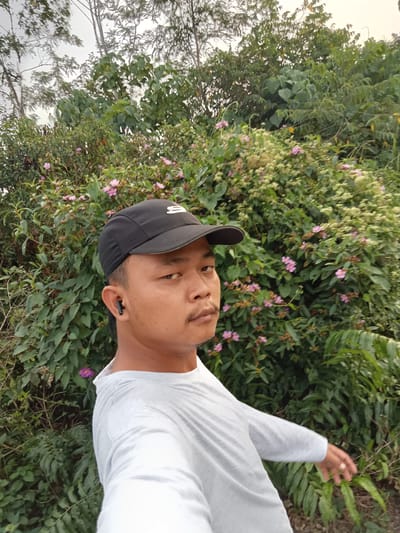 A man wearing a black baseball cap and a light gray long-sleeved shirt is captured in a selfie. He has a mustache and is wearing a black earbud in his left ear. The man is positioned in front of a dense green foliage backdrop with scattered small pink flowers. The scene appears to be outdoors, possibly in a natural or park-like setting. The sky is overcast and light-colored. The man's right arm is extended towards the right side of the frame, with his hand visible. The overall impression is a casual outdoor portrait. The location is identified as Teluk Kenidai, Indonesia.