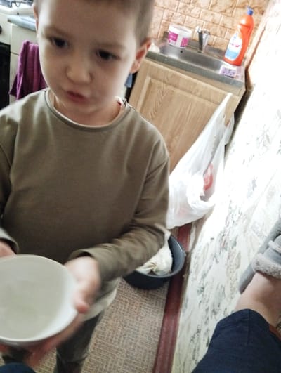 A young boy, appearing to be of Caucasian ethnicity and approximately 4-6 years old, is the central subject of the image. He is wearing a long-sleeved, olive-green shirt and dark pants. The boy is holding a white bowl in his hands, angled towards the viewer, suggesting he might be interacting with it, perhaps about to eat or drink from it. His facial expression is somewhat serious, with his mouth slightly open.

The background of the image reveals a domestic environment, likely a kitchen or bathroom area, given the presence of a sink with a faucet, a wooden cabinet, and cleaning supplies on the countertop. Specifically, an orange bottle of liquid, possibly dish soap or a cleaning agent, and a white container with a pink lid are visible. A white plastic bag hangs near the sink. The walls are tiled with a beige, brick-like pattern. The floor appears to be carpeted with a patterned texture.

The overall lighting suggests an indoor setting, possibly with artificial light sources. The image composition is a medium shot, focusing on the boy and the immediate objects he is interacting with, while also providing context of his surroundings. The scene does not contain any elements that specifically identify its location within Kingisepp, Russia.
