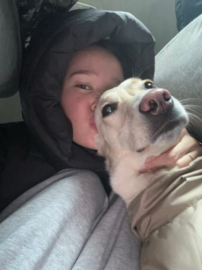 A medium close-up shot captures a person and a dog within an indoor setting at Yemelyanovo Airport, Russia. The human subject, a female, is positioned on the left, wearing a black hooded puffer jacket with the hood obscuring much of her head. Her face, featuring light skin and dark eyes, is visible from the eyes down, directed slightly right of the camera with lips pursed in a kissing gesture. She is also wearing grey sweatpants. Her left hand, adorned with a thin gold ring, supports the neck of a light-colored dog. The dog, appearing cream or light tan, occupies the right side of the frame. It wears a light brown or tan coat, has a dark pinkish nose, and its dark eyes are directed upwards and slightly left. Its head is tilted upwards towards the camera. The background consists of indistinct grey fabric, consistent with seating or interior paneling.