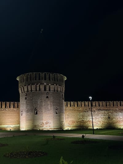 The image shows a night view of a brick fortress wall with a round tower on the left, illuminated by artificial lights. The fortress is located in the city of Smolensk, Russia, not Radkevshchina. The wall has crenellations along the top and small arched windows. The round tower is also made of brick and features similar architectural details. In the foreground, there is a grassy area with a pathway. A single lamppost stands to the right of the tower, casting light on the wall. The sky is dark, indicating nighttime. There are no people or animals visible in the image.