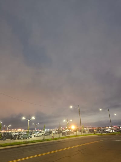 The image depicts a parking lot at night in Dorval, Canada, likely near the airport. The sky is overcast with diffused light, suggesting twilight or a cloudy evening. Numerous cars are parked in rows under the illumination of streetlights. A chain-link fence separates the parking lot from a road in the foreground. In the distance, city lights and airport infrastructure are visible. There are no people or animals present in the frame.