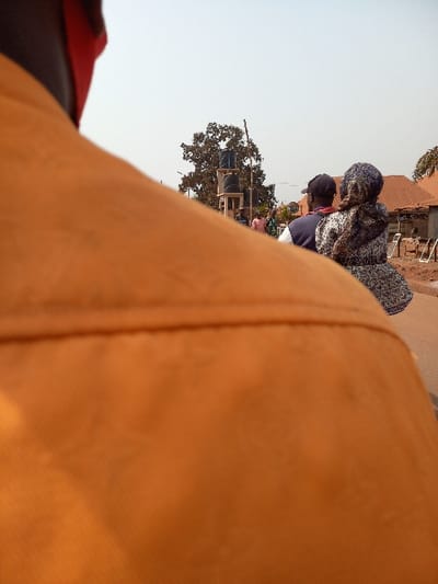 This image captures a street scene in Oturkpo, Nigeria, observed from an over-the-shoulder perspective. In the immediate foreground, the upper back and shoulder of an individual wearing a solid orange garment are visible, with a portion of a red face covering near the left ear. This foreground element is slightly out of focus.
In the midground, two individuals are seated on what appears to be a motorcycle or scooter, moving away from the viewer on a light-colored road surface. The driver is a male, wearing a dark baseball cap and a dark blue sleeveless vest over a lighter shirt. Seated directly behind him is a female passenger, wearing a patterned dress with a distinct dark and light motif, and a matching or complementary patterned head covering. Her right arm is visible, resting on the driver's back.
The background features a mix of natural and man-made elements under a clear, bright sky. To the left, a large tree with dense, dark green foliage is prominent. Beneath the tree, a structure resembling a water supply point is visible, featuring stacked dark blue plastic water tanks on a multi-tiered stand. Further to the right in the background, several single-story buildings with reddish-brown corrugated metal roofs are present. In front of one building, multiple white plastic chairs are arranged along the roadside. The ground surface generally appears light brown, consistent with an unpaved or dusty environment. Distant figures and faint structures can be discerned along the street.