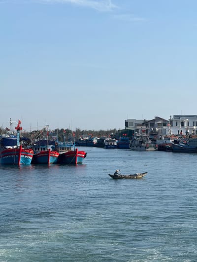 The image shows a waterfront scene in the city of An Hải, Vietnam. In the foreground, the blue water of a river or canal is visible with some white foam at the bottom right, suggesting a wake or current. A small, wooden boat with a single occupant wearing a conical hat is positioned in the middle of the water, facing towards the left.

In the middle ground, several large, colorful fishing boats are docked side-by-side. These boats are predominantly red and blue, with red flags flying from their masts. Numbers and text are visible on the hulls of some of the boats, such as "Q.N. 91233.BL" and "Q.N. 91244.BL".

In the background, a line of buildings borders the waterfront. These include a white, multi-story building with ornate architectural details, a darker, more industrial-looking building, and other structures. Trees are visible behind the buildings, and the sky above is clear and blue with a few wispy clouds. The overall impression is of a bustling harbor or port area.