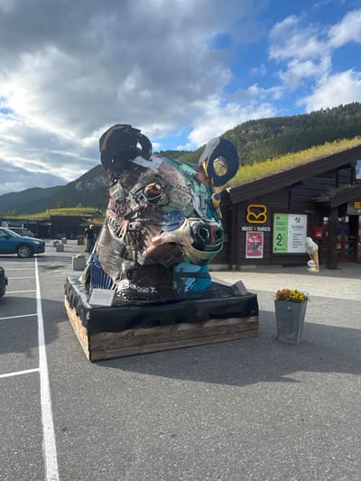 In Røkland, Norway, a large, abstract sculpture of a bear's head, constructed from scrap metal and repurposed materials, stands prominently in an outdoor parking lot. The sculpture is situated in front of a building with a distinctive green, grass-covered roof, suggesting a natural or eco-friendly architectural design. Behind the sculpture, a building with signage that includes "HOUSE OF BURGER" and "SALTDAL" is visible, indicating it's likely a commercial establishment, possibly a restaurant or service area. The sky above is partly cloudy, with patches of blue sky and scattered white clouds. In the background, rolling hills covered with trees and vegetation are visible. The parking lot itself is paved and marked with white lines. Several objects are scattered around the scene, including a blue car parked on the left, a planter with flowers in the mid-ground, and a large cutout advertisement of an ice cream cone. A person is walking in the background near the building. The sculpture is placed on a wooden base covered with black material.