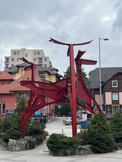 The image displays a prominent, large, abstract red metal sculpture, approximately 5-6 meters in height, situated on a grey paver pedestrian area in the town of Zlatibor, Cajetina Municipality, Serbia. The sculpture consists of intersecting flat and curved steel panels, forming a stylized, multi-limbed figure. Surrounding its base are stone-bordered planters containing dense evergreen shrubs.
In the midground, a paved road or driveway descends, with a silver sedan and other parked vehicles visible. To the left of the road, a reddish-orange building with a red tiled roof and balconies features a partially visible sign reading "ЗЛАТИ СПЕЦИЈАЛ." Behind this, a yellow building with a brown tiled roof is present. In the upper left background, a modern, multi-story building with a light-colored facade and numerous rectangular windows rises above the surrounding structures. An antenna or communication tower is visible adjacent to this building. To the right of the road, a brown wooden-clad building with a dark roof and lighter sections is visible. A tall grey street light pole stands to the right of the sculpture. Blue refuse bins are located near the descending road. The sky is overcast and grey. No individuals are visible in the scene.