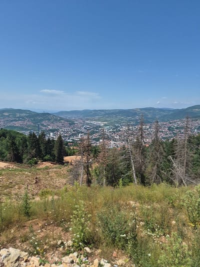 The image depicts a panoramic view of a city nestled in a valley, with rolling hills in the background under a clear blue sky. In the foreground, there is a clearing with dry, bare trees and sparse vegetation. The city itself is densely populated with buildings of varying sizes and colors, many with red-tiled roofs. Patches of green trees are scattered throughout the urban landscape and on the surrounding hillsides. There is evidence of construction or development in the foreground, with disturbed earth and some construction machinery visible. The overall scene suggests a large urban area surrounded by nature. Based on the context provided, this is a view overlooking the city of Sarajevo, specifically from the area of MZ Trebević, Bosnia and Herzegovina, a location known for its elevated position offering city views.