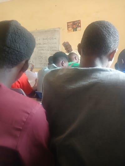 The scene depicts an indoor classroom in Ugi Majowel, Nigeria. The perspective is from behind two individuals in the foreground, looking towards the front of the room. The individual on the left wears a dark red long-sleeved shirt, and the individual on the right wears a dark olive green short-sleeved top. Both individuals have short, dark, curly hair.
In the midground, multiple other individuals, primarily visible from the back of their heads and shoulders, are seated at desks facing forward. One of these individuals wears a green shirt. At the front of the room, a bearded male, identified as a teacher, stands facing the seated students. He wears a patterned traditional cap. Behind the teacher, a white board displays handwritten mathematical equations and expressions in black marker, including "3mt + (T-t)dxx" and "Tc = 180". A colorful poster featuring human figures is affixed to the light yellow wall to the right of the whiteboard. Papers are visible on some desks in front of the seated students.
