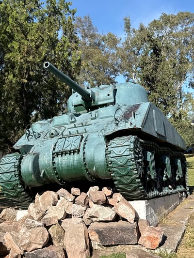 A World War II tank, painted green and surrounded by rocks, is positioned outdoors in Salta, Argentina, with trees and a blue sky in the background.