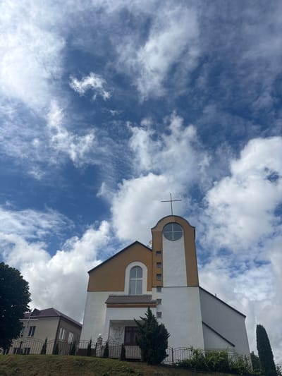 An exterior, low-angle, eye-level, full shot captures a modern church building with a tall, slender steeple topped with a cross, set against a vibrant blue sky filled with scattered white clouds. The church features a white lower facade, a tan upper section, and a distinctive arched window with a circular pane on the steeple. To the left of the main structure, a smaller, lighter-colored building with a dark red roof is visible. The church is situated on a grassy slope, bordered by a dark metal fence and green bushes. A tall, dark green evergreen tree stands to the right of the church. The scene appears to be located in Капыльскі сельскі Савет, Belarus, given the context. There are no people or animals visible in the image.
