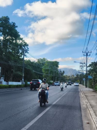 A person is riding a motorcycle on a paved road in Baan Thong Po, Thailand. The person is wearing a helmet and a light-colored shirt and blue jeans. They are in the foreground, and the motorcycle is facing away from the camera. The road is two lanes wide, and there are other vehicles on the road, including a black SUV, a white car, and two other motorcycles. There are trees and power lines on both sides of the road. In the background, there are mountains. The sky is blue with some white clouds. The scene appears to be a typical street view in a tropical location.