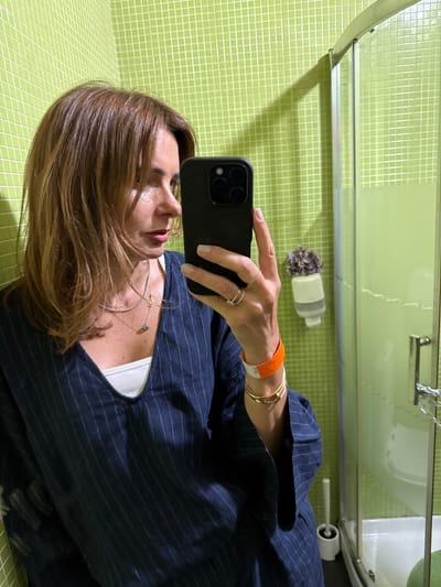 A woman with brown hair and wearing a dark blue pinstripe top is taking a mirror selfie in a bathroom. The walls of the bathroom are covered in light green square tiles. To the right of the woman, a glass shower enclosure is visible. The woman is holding a black smartphone in her right hand, and is wearing several rings on her fingers, as well as necklaces and bracelets. An orange wristband is also visible on her right wrist. The bathroom also contains a white toilet brush holder with a brush.