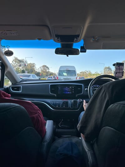 The image depicts the interior perspective from the rear passenger seat of a right-hand drive vehicle, stationary in traffic on a multi-lane road in Narre Warren, Australia. The time is daylight with a clear blue sky.
In the foreground, the backs of the driver's and front passenger's seats are visible. The driver, wearing a dark shirt and blue jeans, is positioned on the right, with their right hand resting near the electronic gear shifter and push-button start on the center console. A phone mount with a navigation application is affixed to the dashboard on the driver's side. The front passenger's seat on the left has a red garment draped over it. A blue baseball cap rests on the floor between the front seats.
The vehicle's dashboard features an infotainment screen displaying "5:19" and music information, positioned above a climate control panel. Charging cables are visible in a storage compartment below the console.
Through the windshield, a queue of vehicles is stopped at a traffic light, which appears red in the distance. A white cargo van is directly ahead in the same lane. Other vehicles (silver, red, dark colored) are visible in adjacent lanes. A round "CPV" (Commercial Passenger Vehicle) tag hangs from the rearview mirror assembly on the passenger side. A dashcam is mounted to the right of the rearview mirror. Trees and general vegetation line both sides of the road in the background. The precise intersection in Narre Warren cannot be determined from the available visual information.