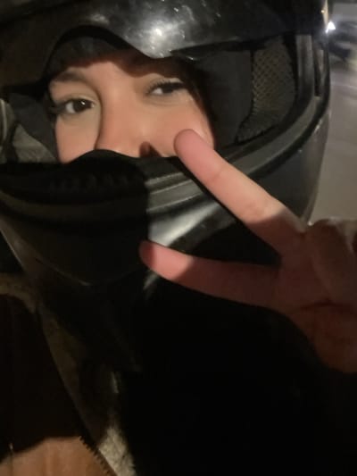 A close-up image depicts an individual in Mechouar Fes Jdid, Morocco, wearing a black full-face motorcycle helmet with the visor open or removed, revealing only their eyes and the bridge of their nose. The person's right hand is raised, forming a "V" or "peace" sign with the index and middle fingers extended, while the other fingers are bent. The visible facial features include brown eyes with distinct eyelashes and eyebrows. The helmet's interior lining, appearing as a dark mesh material, is visible near the left side of the face. The scene is dimly lit, suggesting night or low-light conditions, with a blurred, dark background. A portion of dark outerwear with a lighter, textured lining is visible in the lower left foreground.