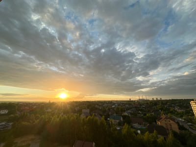 This is an aerial, wide-angle, eye-level shot of a semi-rural landscape in the vicinity of Schomyslicki Sel'ski Sovet, Belarus, during a sunset. The sun, a bright orb on the left side of the horizon, casts a warm, golden glow across the sky and illuminates the scene. The sky is filled with a mixture of dramatic, dark grey clouds and patches of lighter, more ethereal clouds, with a hint of blue peeking through.
In the foreground, dense green trees dominate the lower portion of the image, with their foliage creating a lush, textured canopy. Beyond the trees, a residential area is visible, characterized by a collection of houses with varying roof colors, including red, brown, and dark grey. Some houses have visible windows and architectural details. The streets are lined with parked cars, suggesting an inhabited area.
In the distance, several industrial structures, likely power plant cooling towers, stand against the skyline, emitting plumes of steam or smoke. The overall environment appears to be a blend of natural greenery and human development. The lighting conditions indicate either early morning or late evening due to the low angle of the sun and the warm hues. No people are visible in the image.