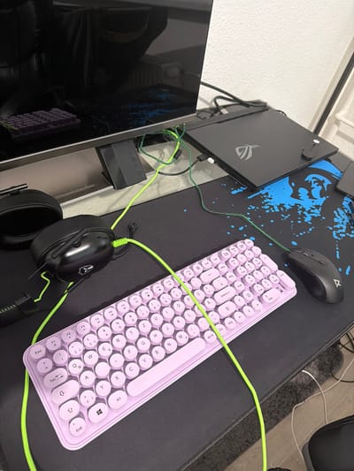 The image shows a gaming setup. In the foreground, a light purple, compact keyboard with round keys is placed on a black mousepad. A black gaming mouse is positioned to the right of the keyboard. To the left of the keyboard are black headphones with neon green accents. A neon green cable runs from the headphones and connects to something out of frame. In the background, a black laptop with a silver logo sits on a desk. The screen of a monitor is visible, reflecting purple keys. Wires, predominantly neon green, are neatly organized. The environment appears to be a home office or gaming station. The image was taken in Halle (Saale), Germany.