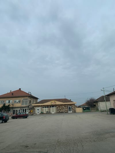 The scene depicts an outdoor view in the city of Kozar Belene, Bulgaria, under an overcast sky. The foreground comprises a large, uneven asphalt or paved surface with visible cracks. In the midground, on the left, stands a two-story building with a light beige facade and a red-tiled hip roof. Its ground floor features multiple windows and entrances, and several external air conditioning units are affixed to its exterior. A dark-colored SUV is partially visible parked on the far left, followed by a red station wagon parked in front of the two-story building. To the right of this building, a single-story structure with a similar red-tiled hip roof is visible. Its facade features a light-colored frieze with a central circular emblem depicting figures, and the lower portion is adorned with irregular, light brown stone or tile work. Multiple large, white-framed window openings are present, some appearing covered from within. A bicycle is leaning against the front of this building. Overhead power lines span across the midground. To the right, a green metal gate and a light-colored residential building are partially visible, along with a black wheeled trash bin and a street light pole on the far right. The background shows sparse, bare trees. No individuals are present in the scene.