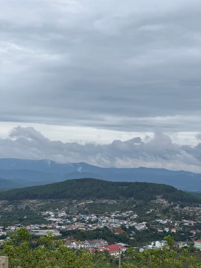 The image depicts a panoramic view of Trường Thọ, Vietnam, on an overcast day. The left portion of the frame is dominated by a gray, cloudy sky, transitioning to a thinner, brighter band of clouds closer to the center. In the middle ground, a series of forested mountains gradually fade into the distance with a blueish hue. The right side of the image shows a dense collection of buildings, likely homes and businesses, clustered along the slopes of a tree-covered hill. The buildings are of various colors and sizes, with red roofs visible among them. A cluster of green foliage occupies the extreme right edge of the frame, obscuring a portion of the urban landscape.