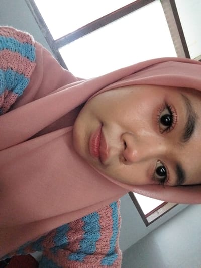 A young woman, wearing a pink hijab and a pink and blue striped sweater, looks up at the camera. She is indoors, with what appears to be a window or skylight in the background. The image is a close-up of her face, with her eyes, nose, and mouth visible. Her makeup includes mascara, eyeliner, and lip gloss. The overall lighting is bright, suggesting natural light from the window. There is no indication of the specific location within Tibojong, Indonesia.